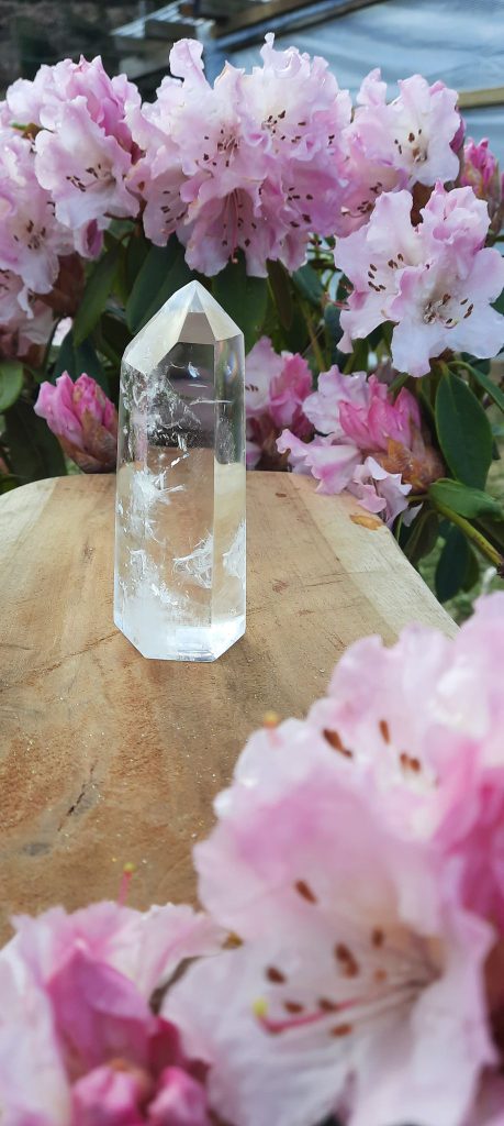 Cristal de roche taillé en obélisque posé sur une table entouré de fleurs roses.